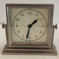 Photo of silver metal dresser clock showing front face with black Arabic numerals and black hour and minute hands on white background, mounted on a decorative metal base.