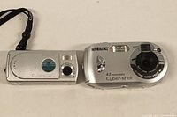 Two silver Sony Cyber-shot digital cameras side by side on a white surface showing front features and size comparison