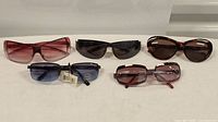 Five pairs of sunglasses on a white surface including various frame shapes and lens colors.