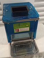 Front view of the meat slicer with blue casing, black hopper on top and stainless steel tray at bottom.