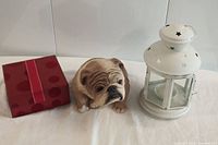 Photo showing ceramic bulldog figurine alongside white metal lantern with star cutouts on a white surface.
