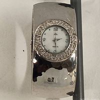 Silver-tone wide bangle watch with rhinestone bezel and white dial showing numeric and dot markers for hours.