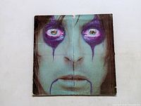 Front cover of Alice Cooper LP showing a close-up face with painted makeup, primarily green and purple, indicating some wear and creases.