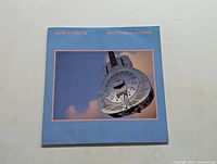 Front cover of Brothers in Arms vinyl LP by Dire Straits showing resonator guitar against blue sky background.