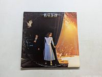 Front cover of Rush 'Exit...Stage Left' album showing a stage exit scene with a woman in a dress and a man behind a curtain with audience and stage lights.