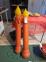 Full view of a pair of tall blow mold candles standing on a floor in front of a red tablecloth-covered table. The candles are orange with yellow flame tops and have electrical cords.