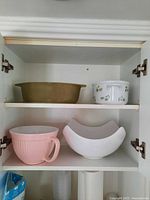 Wooden cabinet shelf with a brown ceramic casserole pot on the top shelf, white ceramic bowl with floral design next to it. Below shelf holds a large pink plastic mixing bowl and a white ceramic curved bowl.