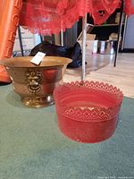 Overall view of brass pot with lion head handles and red decorative metal pot on a carpeted floor.