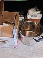 Overview showing cardboard boxes containing glass canning jars and a large stainless steel pot with black handles surrounded by boxed jar lids.