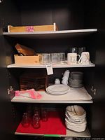 Three shelves inside a dark cabinet with various dishes, glasses, mugs, and folded napkins arranged neatly.