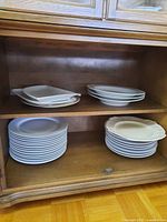 Shelves inside cabinet showing stacked white Ikea plates including both round dinner plates, pasta dishes, and square serving platters