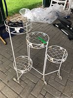 Photo showing front view of the white metal plant stand with four circular tiers arranged at different heights, placed on a paved surface outdoors.