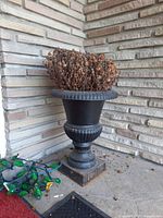 Heavy black metal urn with dried plant inside sitting in a corner outside a building. Shows wear and rust on base. No lights included though green and yellow string lights are visible nearby.