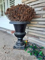 Full view of black metal urn with dried plant arrangement and string lights on ground