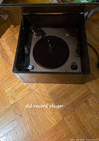 Photo of vintage record player in a wooden cabinet with lid open showing turntable, tonearm, and knobs on parquet wood floor.