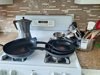 Three Teflon frying pans stacked on a stovetop with an aluminum stovetop espresso maker and kitchen utensils in a metal container.