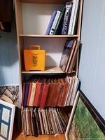 Wooden shelving unit with multiple rows of books and vinyl records. Books include thick dictionaries and other titles; records have various classical music album covers.