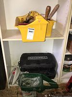 Yellow plastic organizer caddy holding assorted hand tools and wooden handled screwdrivers on a white shelf. Below it a black Rockwell tool case and a green plastic watering can, with a white cordless vacuum on the left side.