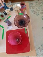 Overview of glass and ceramic vases and bowls arranged on table including pink vase, purple-brown bowl, red square glass bowl, and ceramic floral vase.
