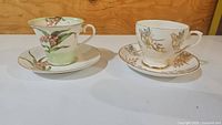 Two vintage teacup and saucer sets displayed side by side on a white surface with a wood panel background, showing colorful floral and gold floral designs.