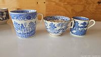 Three antique Blue Willow porcelain cups with two showing visible damage including chips and cracks. The cups feature traditional Blue Willow blue and white transferware design.