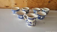 Photo showing six vintage Blue Willow teacups arranged on a white surface with wood background.