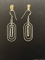 Photo of a pair of .925 sterling silver dangle earrings with elongated oval and paperclip motifs on hook ear wires against a black background.