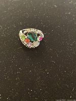 Image showing the front view of a silver-tone ring with a large green teardrop gemstone and multiple smaller colorful stones surrounding it on a black background.