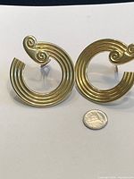 Pair of gold plated swirl hoop earrings with ribbed texture and spiral swirl accent, shown with a coin for size reference.