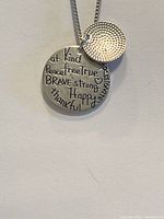 Close-up of the two pendants showing engraved inspirational words on the larger pendant and the word 'be' on the smaller pendant.