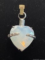 Close-up photo of the heart-shaped moonstone charm showing semi-transparent milky appearance and silver-tone metal prongs holding the stone with a top loop attachment.