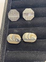 Two pairs of vintage 1970s cufflinks displayed on black padded tray, one octagonal pair with diamond texture and silver stripe, second pair rounded rectangle with gold-tone swirl design.