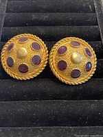 Close-up photo of both round dome-shaped gold plated earrings with purple gemstone accents on a black cushioned display.