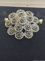 Close-up photo of a vintage filigrana brooch showing green rhinestones forming a floral cluster design with detailed metal openwork.