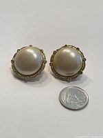 Front view of the pair of vintage clip-on earrings featuring large pearl-like center with small clear crystals around edge and gold-tone metal settings next to a quarter for size reference.