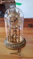Front view of vintage gold-tone anniversary clock with intricate filigree design and Roman numeral face under a glass dome, with winding key included in front.