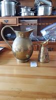 Large rounded brass pitcher with visible patina and wide spout alongside smaller embossed decorative brass pitcher