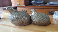 Photo showing two vintage brass quail figurines on a wooden table with detailed feather patterns and natural patina.
