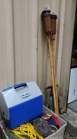Photo of the playmate cooler atop a gray bin filled with camping items and two bamboo tiki torches standing next to it.