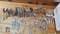 Wide view of all cutlery items laid out on a wooden surface showing knives, forks, spoons in mixed sets and conditions.