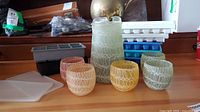 Full view showing glass pitcher, five small swirl pattern glasses in assorted colors, and multiple ice cube trays behind them in various colors and shapes.