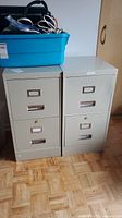 Photo showing two beige metal filing cabinets side by side, both with two drawers each and a blue bin on top with cords.