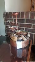 Water fountain on a reflective surface in front of a brick wall, showing multiple cascading tiers with green accents and decorative natural stones in the base.
