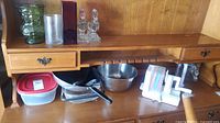 Photo showing various kitchen items including a wok, stainless bowl, plastic lid food container, kitchen slicer with colored inserts, two glass decanters, and horse bookends on a wooden sideboard.