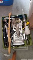 Box with metal graters, wooden spoons, a baster, and various utensils in a green tray.