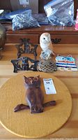 Five owl-themed items on wooden surface: two cast iron trivets, brass trivet, ceramic owl figurine, colorful ceramic trivet.