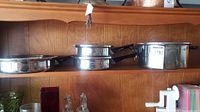 Four stainless steel pots and pans on wooden shelf, showing different sizes and black handles.