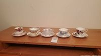 Five assorted fine china teacups and saucers displayed side by side on wooden surface
