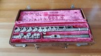 Full view of vintage silver flute disassembled into three parts and placed inside a pink velvet-lined case with brown leather handle.