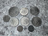 Photo of nine American coins arranged on a stone surface showing both obverse and reverse sides, displaying the Eisenhower dollar, Kennedy half dollars, quarter, steel penny and regular penny.